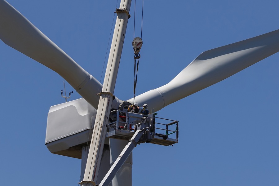 Techniker arbeiten an der Gondel eines Windrades, unterstützt durch einen Kran, unter blauem Himmel.