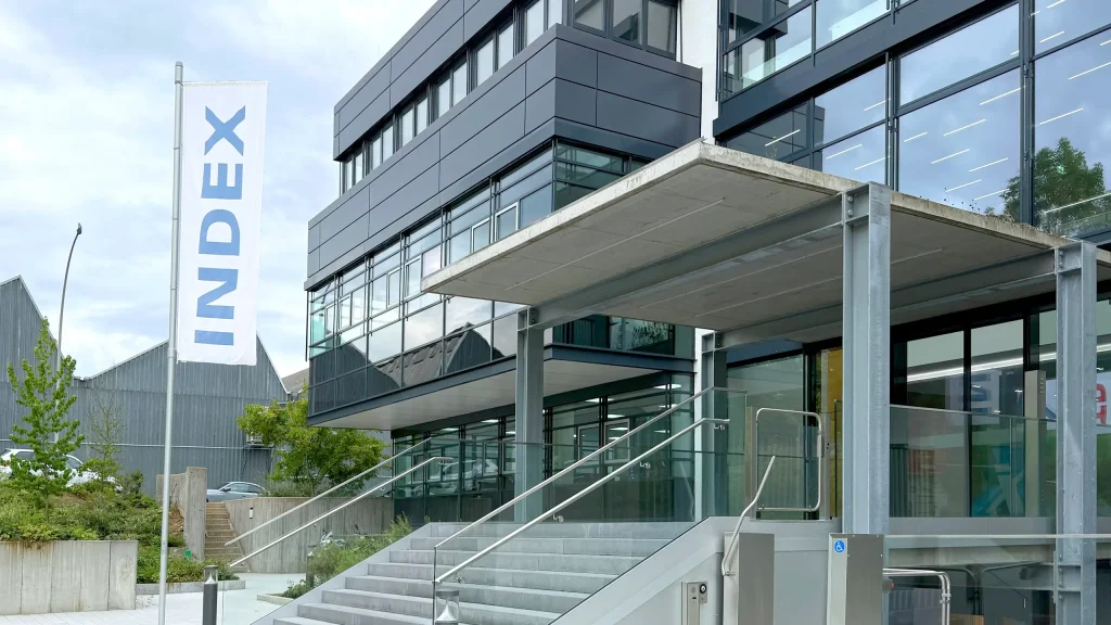 Modernes Bürogebäude mit Glasfassade und Betontreppe. Ein weißes Flaggenbanner mit "INDEX" in blauen Buchstaben weht vor dem mehrgeschossigen Gebäude. Klare, zeitgenössische Architektur mit viel Glas und Stahl.