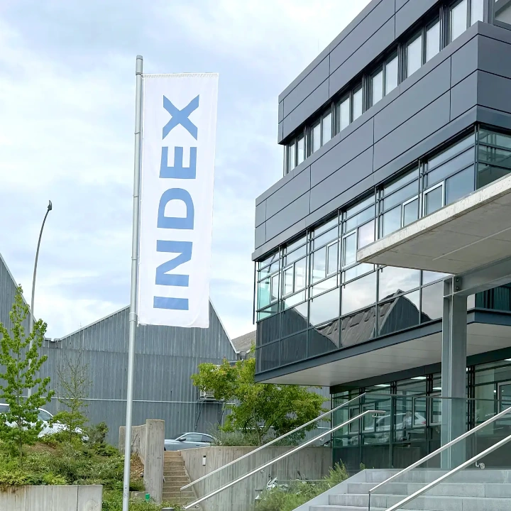 box-id_success-story_index-werke_central Modernes Bürogebäude mit Glasfassade und Betontreppe. Ein weißes Flaggenbanner mit "INDEX" in blauen Buchstaben weht vor dem mehrgeschossigen Gebäude. Klare, zeitgenössische Architektur mit viel Glas und Stahl.